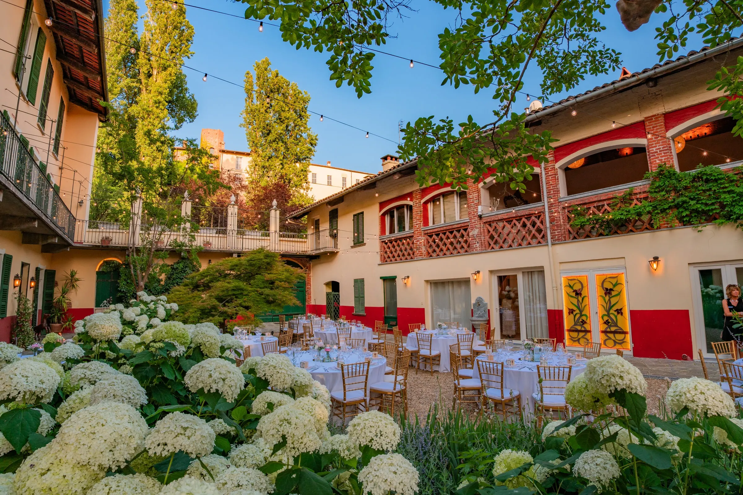 Le ortensie bianche in primo piano e i tavoli rotondi apparecchiati in corte nell’ultima luce del giorno, con la sala superiore accesa dietro le arcate